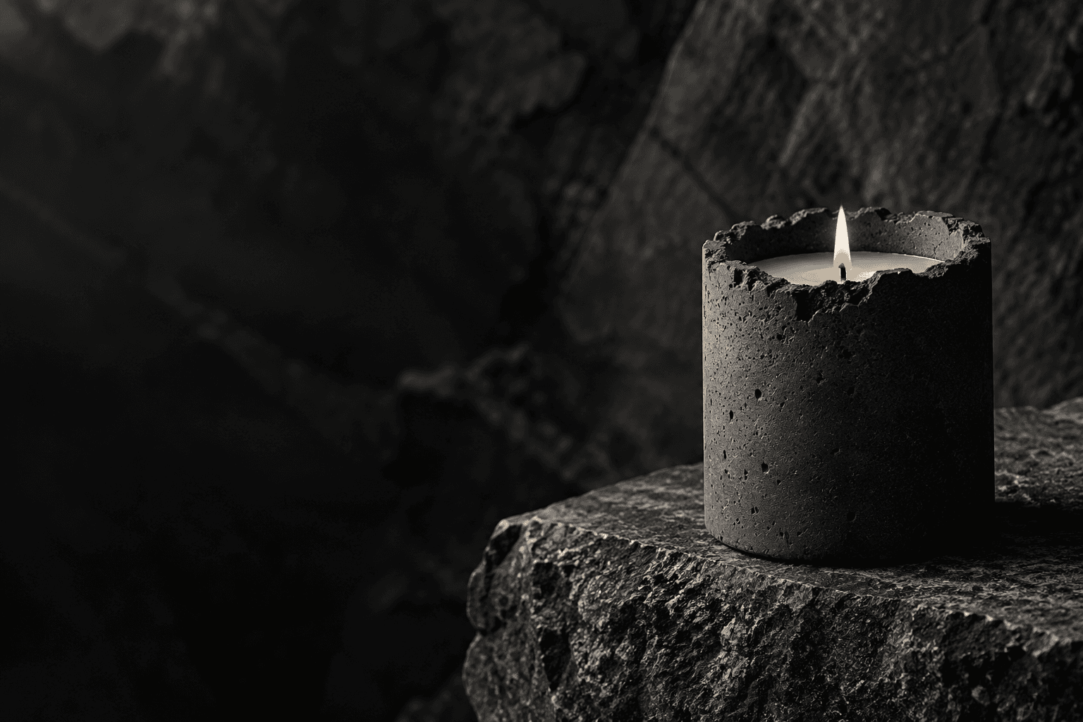 A sculptural charcoal concrete candle with one small flame, resting on raw stone